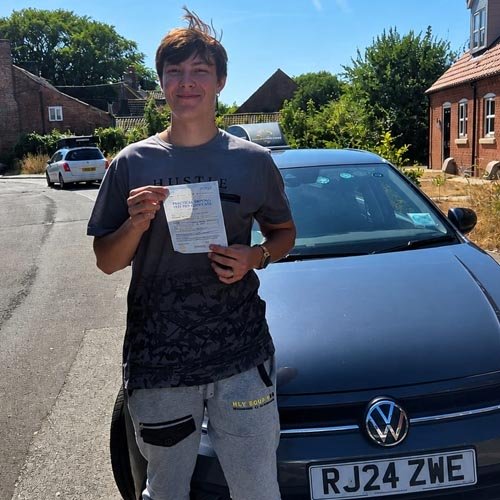 Teenager standing on a sunny suburban street, holding a paper in front of a blue Volkswagen with UK license plate RJ24 ZWE.