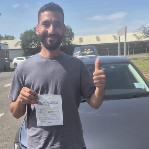 Man with a short beard standing in a parking lot, holding a certificate and giving a thumbs up beside a parked car.
