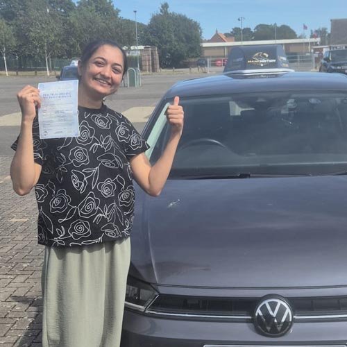 Young woman in a black floral shirt and gray skirt smiles while holding a document and giving a thumbs-up beside a dark VW car in a parking lot.